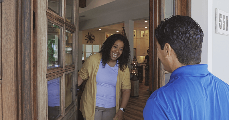 A woman is holding her front door open and smiling at a male Strada Services employee in a blue polo shirt.