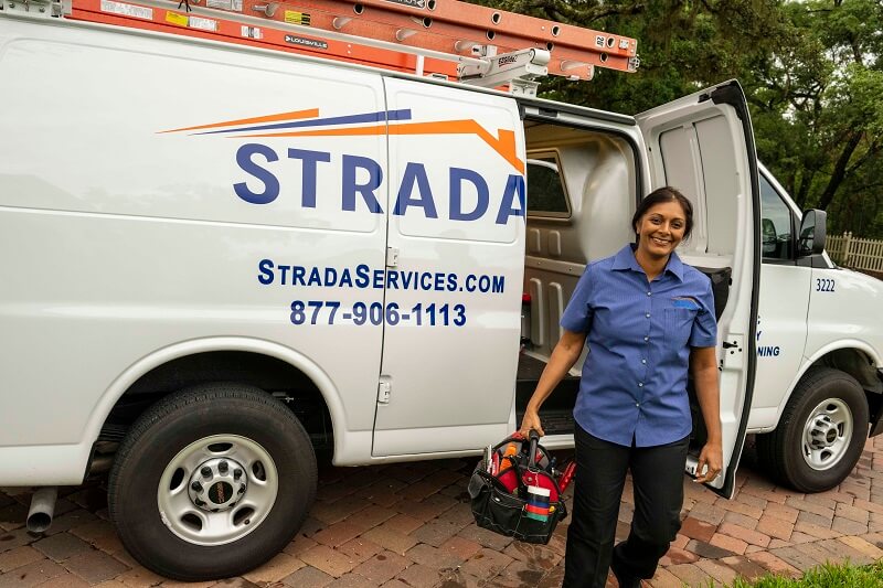 A woman is smiling and carrying a tool bag. Behind her is a white Strada Services utility van with a red ladder on the roof.