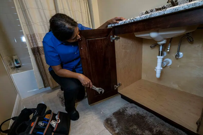 A woman is kneeling down in front of the bottom cabinet of a sink with a wrench in her hand. She's looking at the sink pipes.