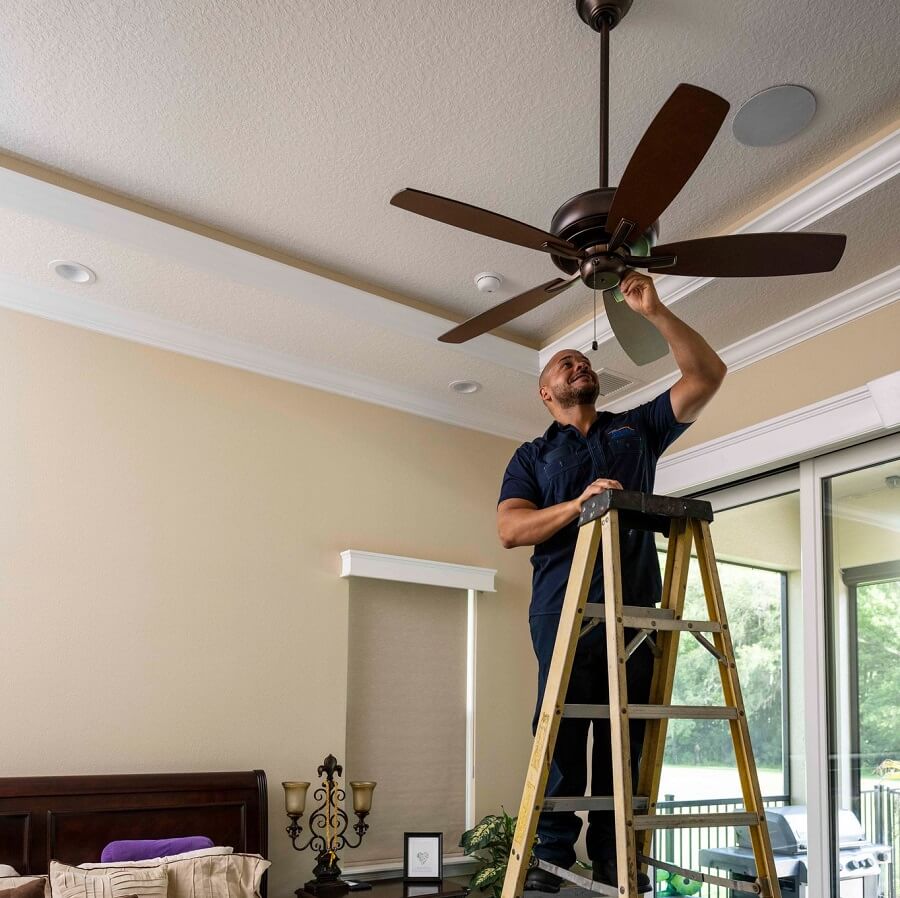 A man is standing on a ladder in a bedroom installing a ceiling fan.