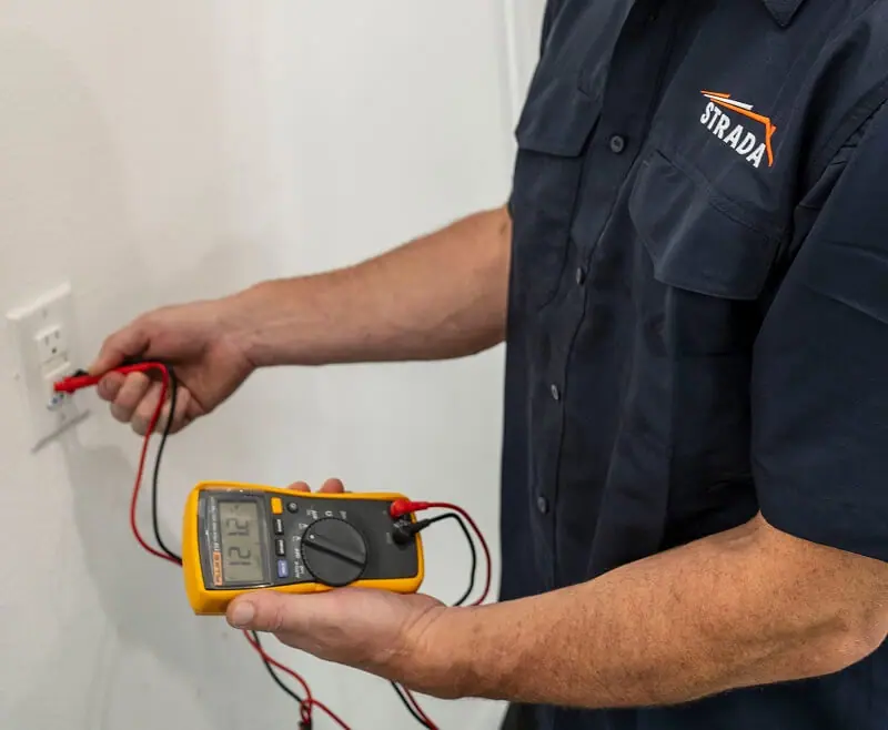 An electrician is holding an electrical device with red and black wires that are plugged into the wall.