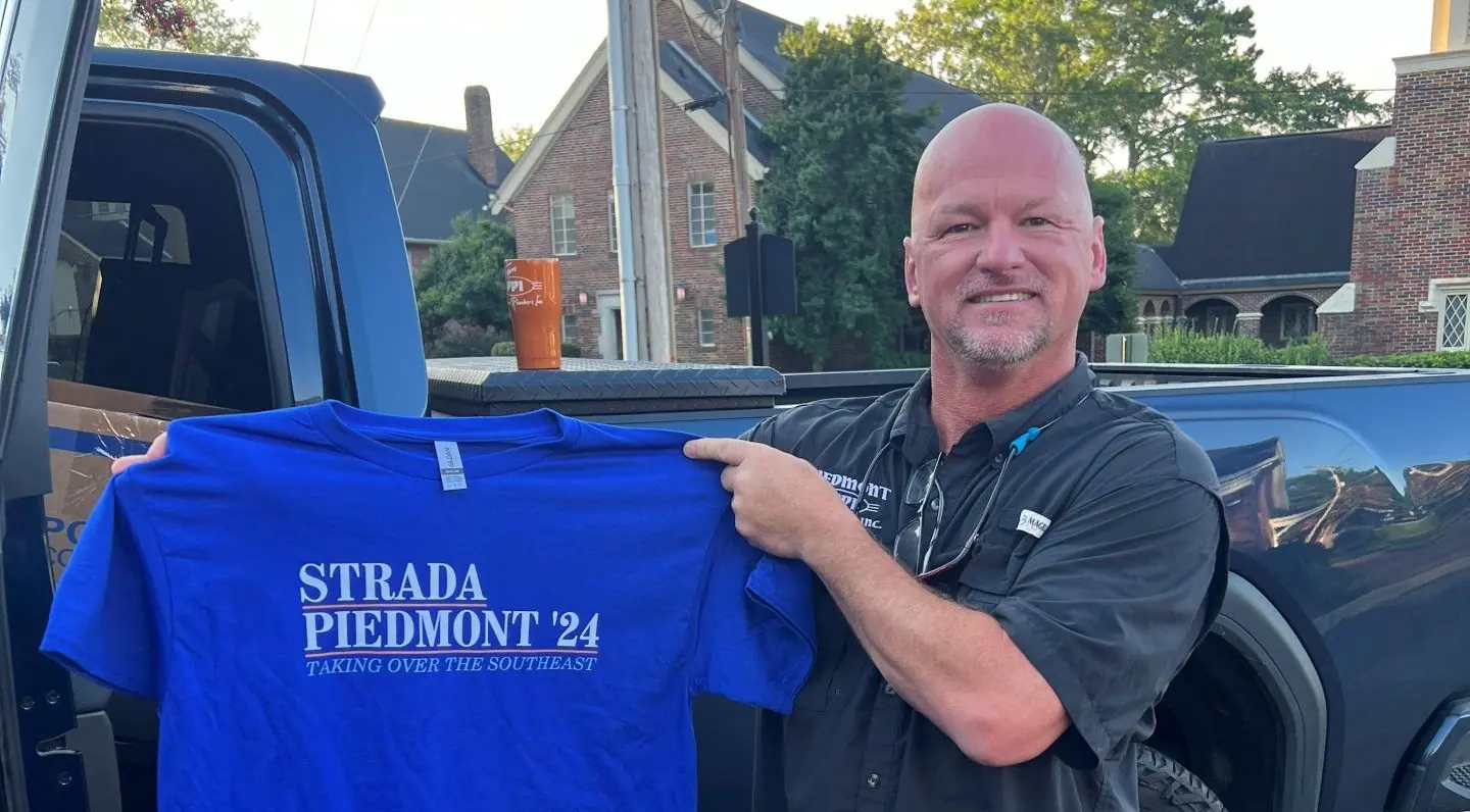 Piedmont Plumbers team lead holding up a blue shirt that says "Strada Piedmont '24"