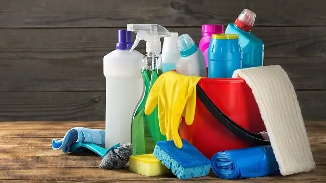 A red bucket sits on top of a wood table top. The bucket is full of cleaning supplies and supplies sit beside it too.